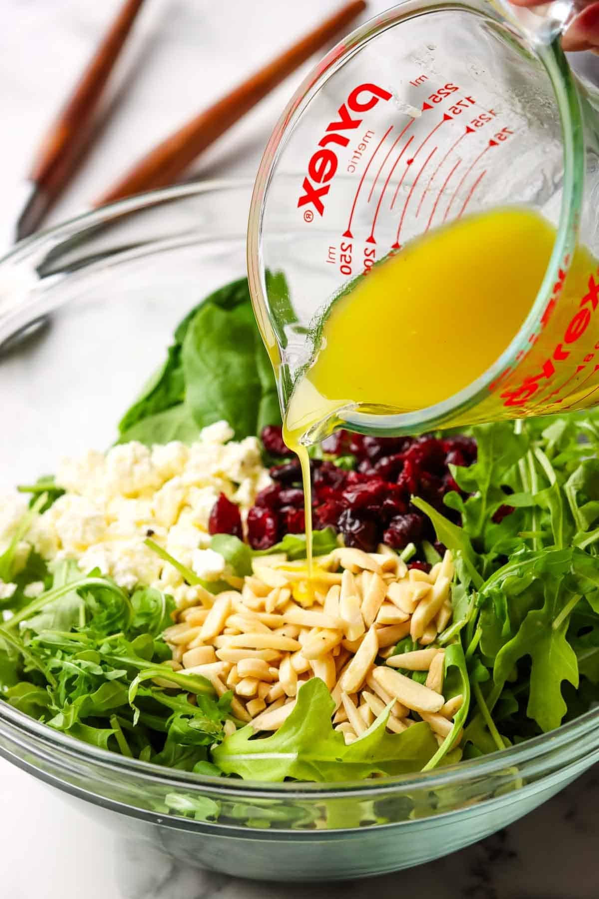 A glass bowl of spinach, arugula, feta, cranberries, almond slices, and more being coated in a homemade vinaigrette salad dressing.