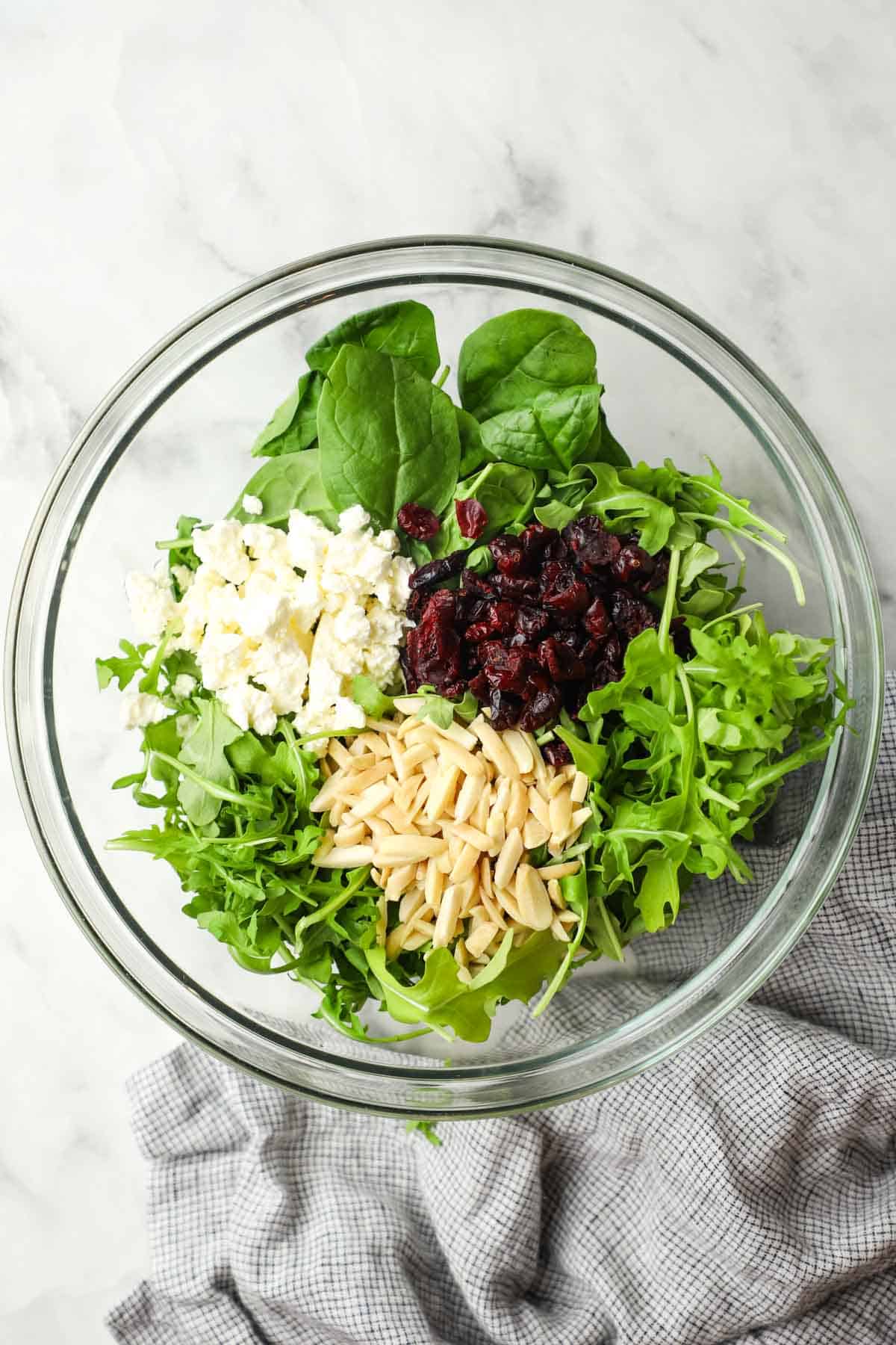 Spinach, arugula, cranberries, feta cheese, and almond slices in a glass bowl.