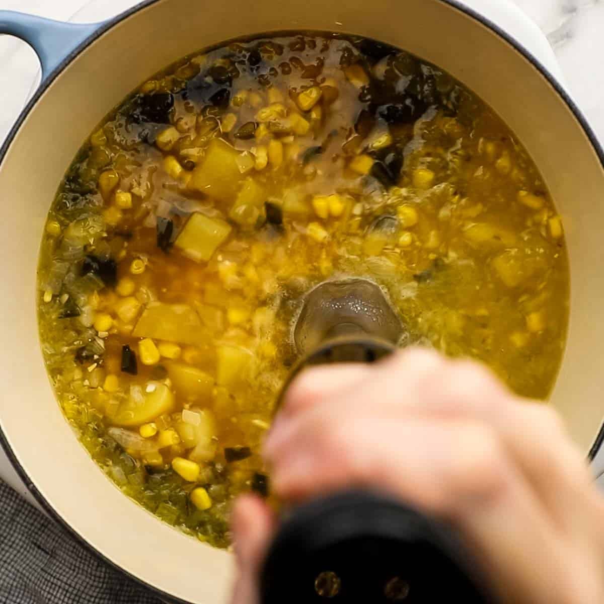Top-down view of immersion blender being used to mix together soup ingredients like poblano peppers, corn, potatoes and more. 