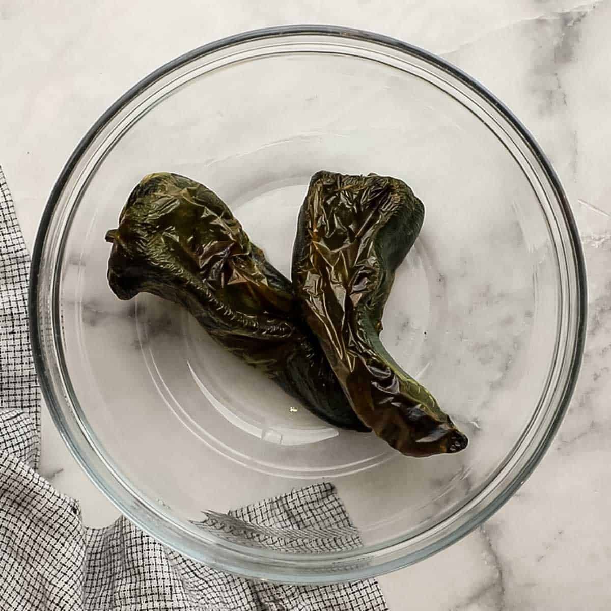 Two poblano peppers that have been roasted in a glass bowl. 