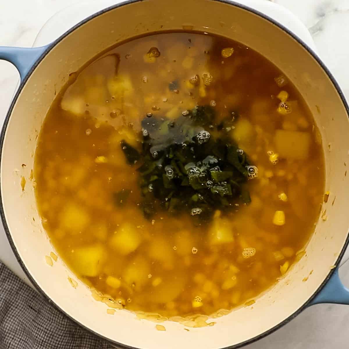 Chicken stock and diced poblano peppers in a soup pot with simmering potatoes, garlic, corn, and olive oil.