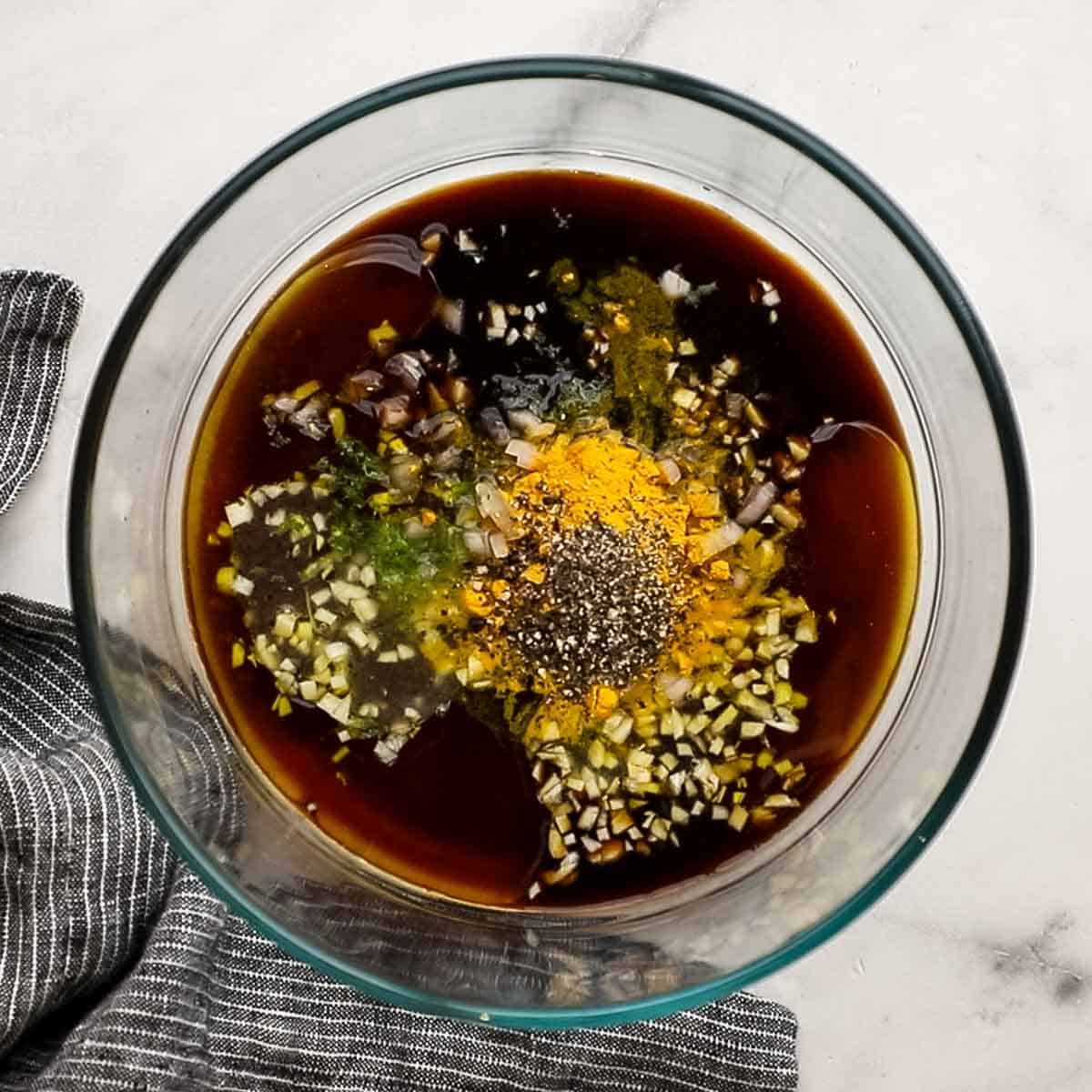 View of ingredients for chicken skewers in a glass bowl that include soy sauce, brown sugar, extra virgin olive oil, fish sauce, garlic, lime zest, lime juice, shallot, turmeric, and black pepper.