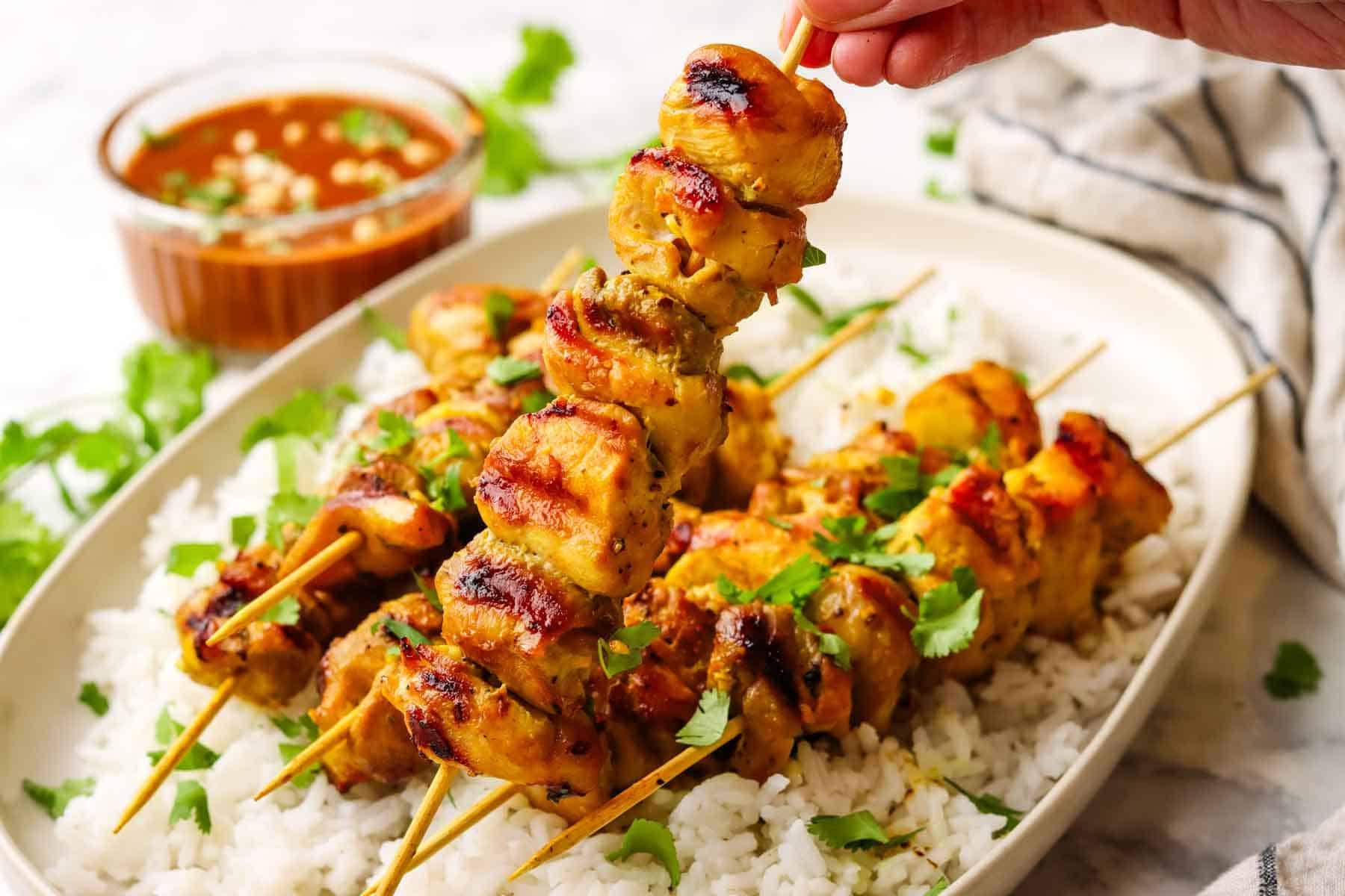 A platter of chicken skewers over rice with a hand lifting one up.