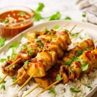A plate of vietnamese chicken skewers on a bed of white rice with a bowl of peanut sauce.