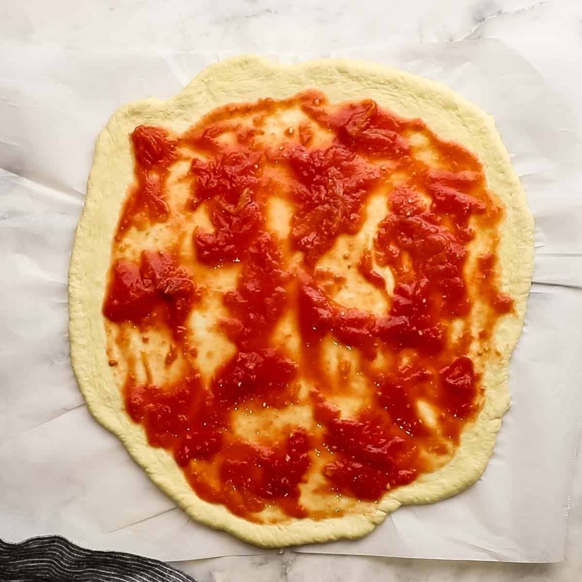 Top-down view of pizza dough that has been rolled out and topped with homemade pizza sauce