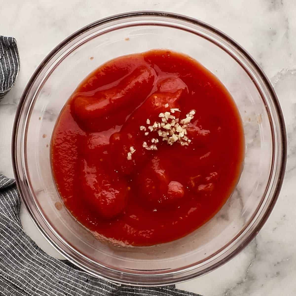 Top down view of pizza sauce ingredients in a bowl that include tomatoes, garlic, and salt.