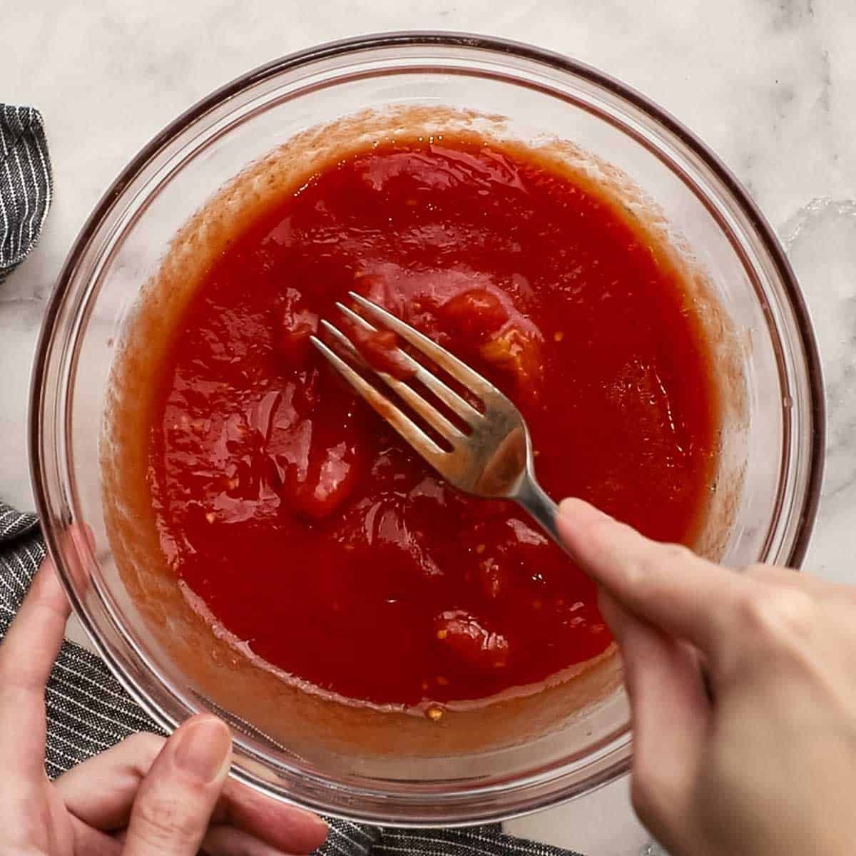 The pizza sauce that contains olive oil, tomatoes, and garlic being mashed together with a fork inside of a glass bowl.