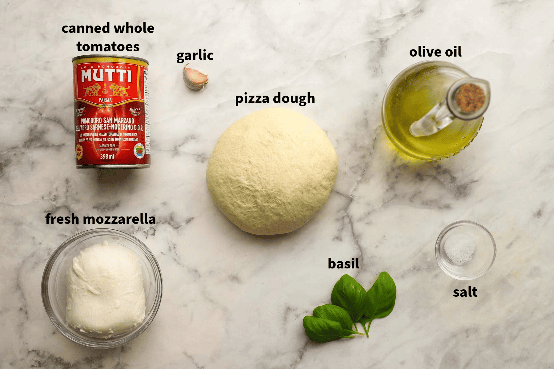 Top-down view of margherita pizza ingredients lying on a counter that includes tomatoes, pizza dough, fresh basil, olive oil, garlic, and more.