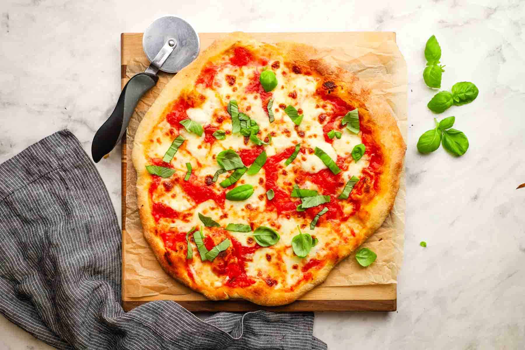 Top-down view of a margherita pizza topped with fresh basil and sitting on a wooden cutting board with a pizza cutter.