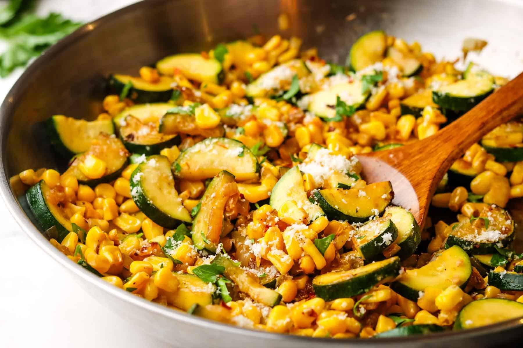 A pain of sauteed corn and zucchini with a serving being scooped out with spoon.