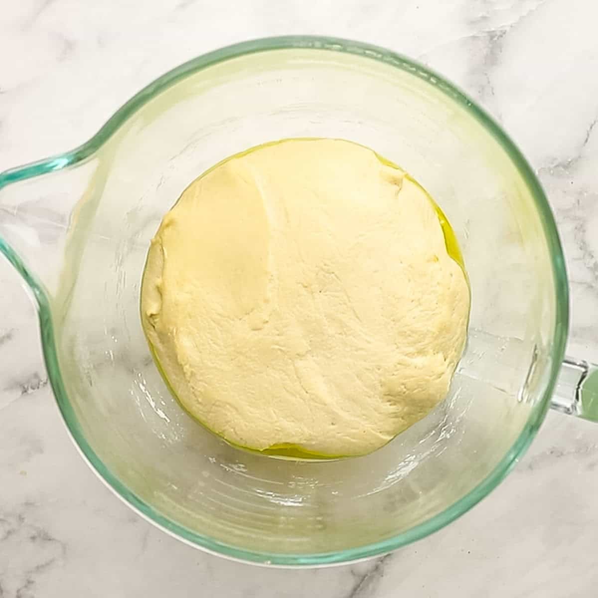 A view of pizza dough rising and resting in a glass bowl.