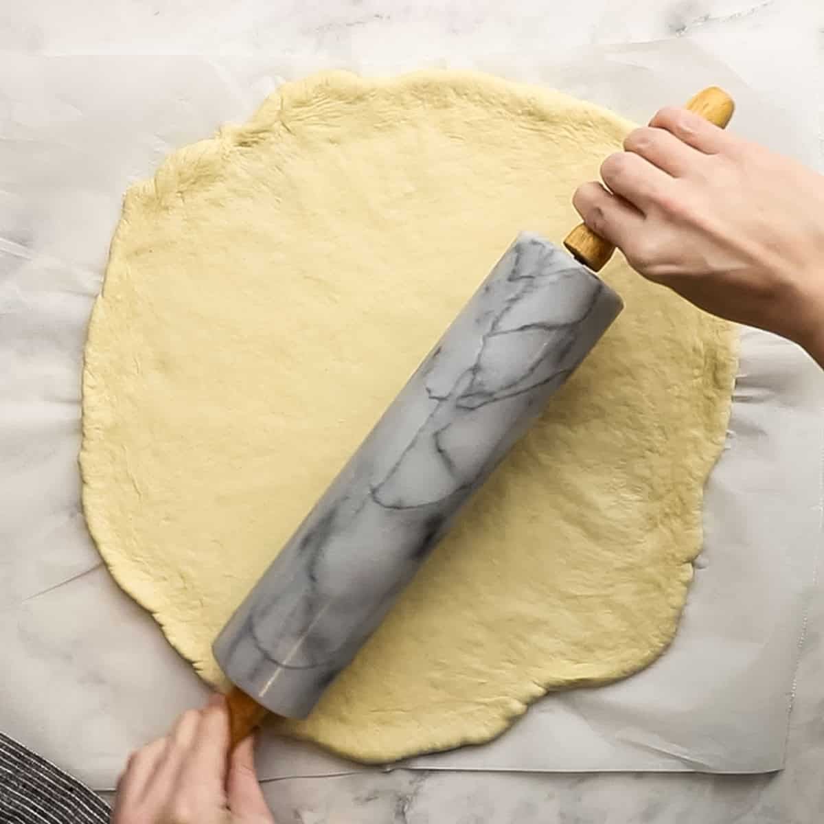Pizza dough being spread thin with a rolling pin on top of parchment paper.