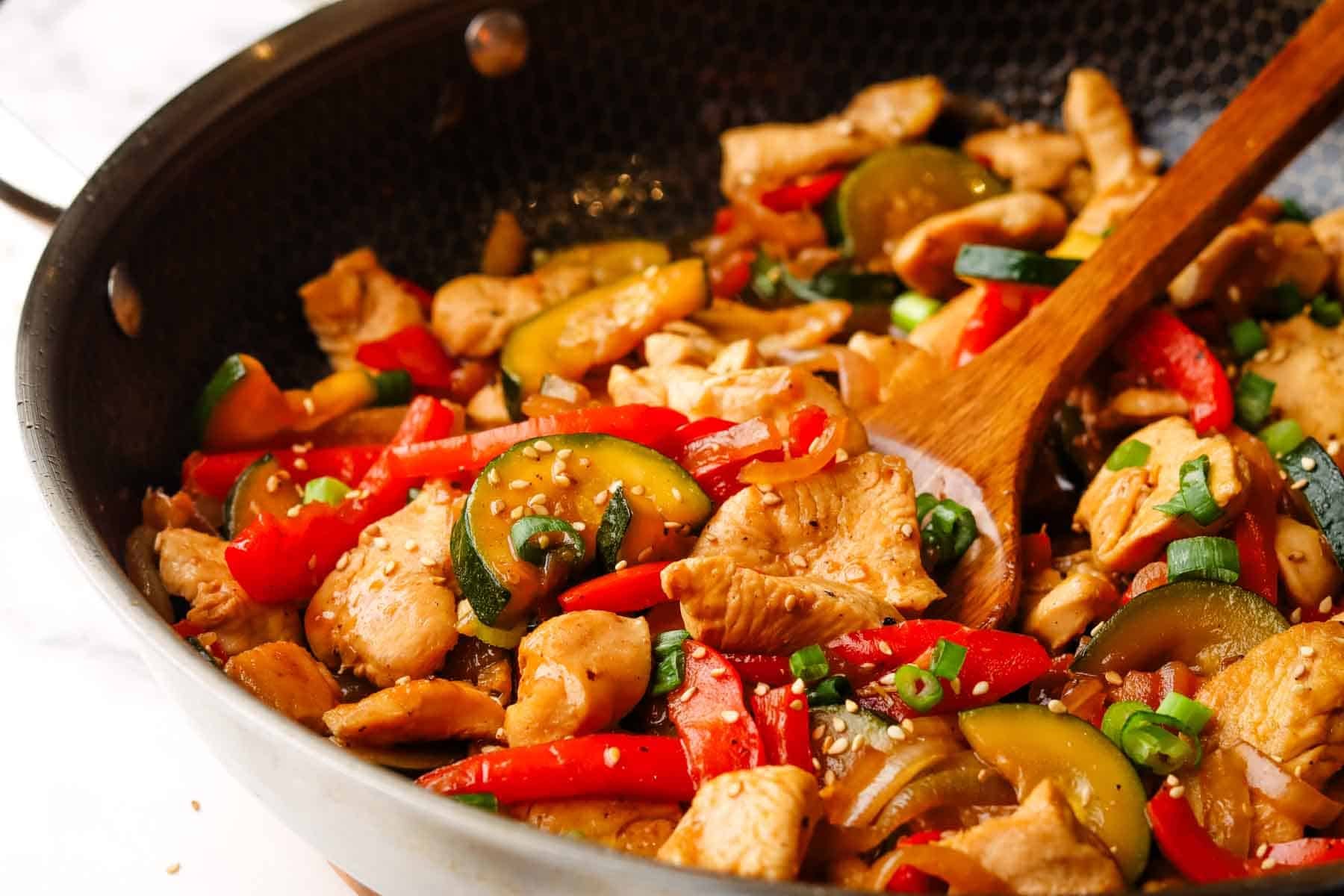A pan of zucchini chicken stir fry with red peppers garnished with sesame seeds.