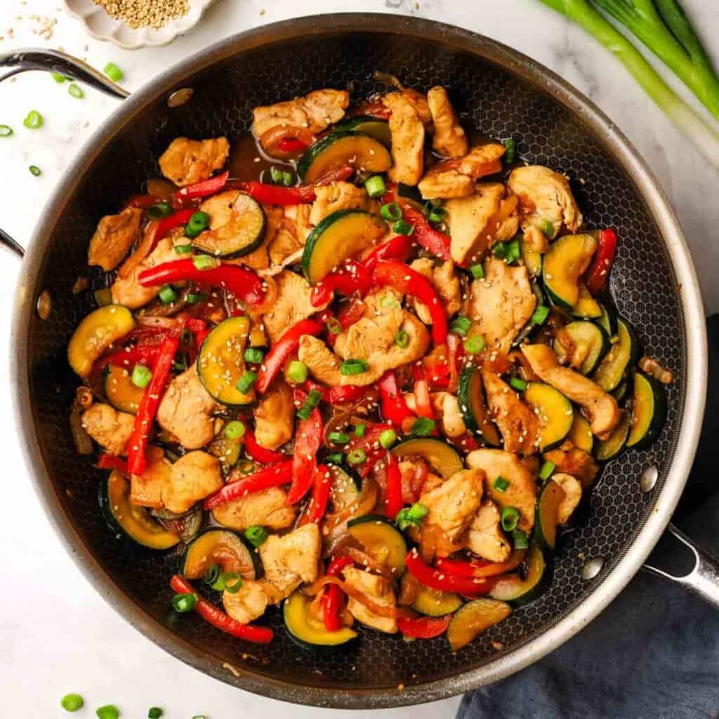 A pan of zucchini chicken stir fry with red bell peppers.