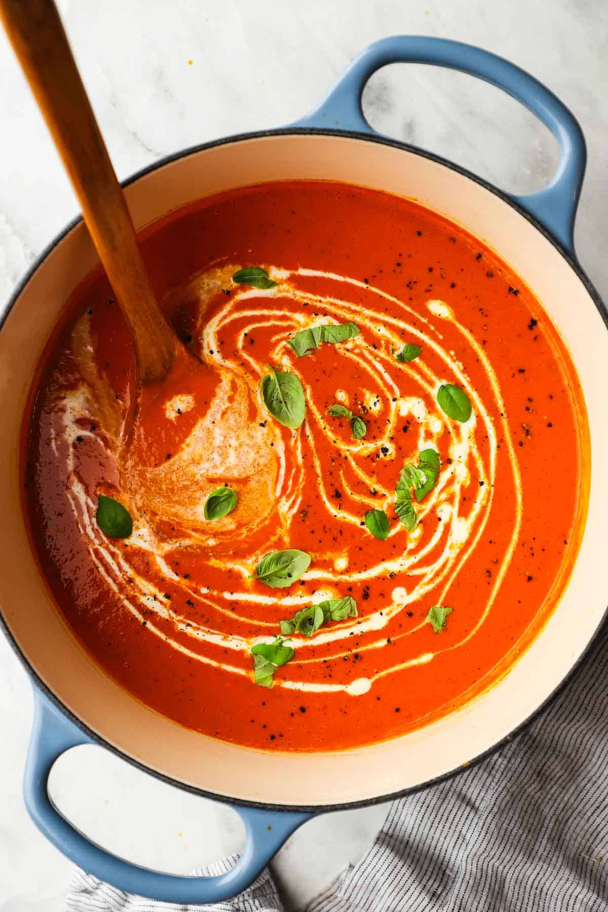 A pot of red pepper soup garnished with cream and fresh basil.