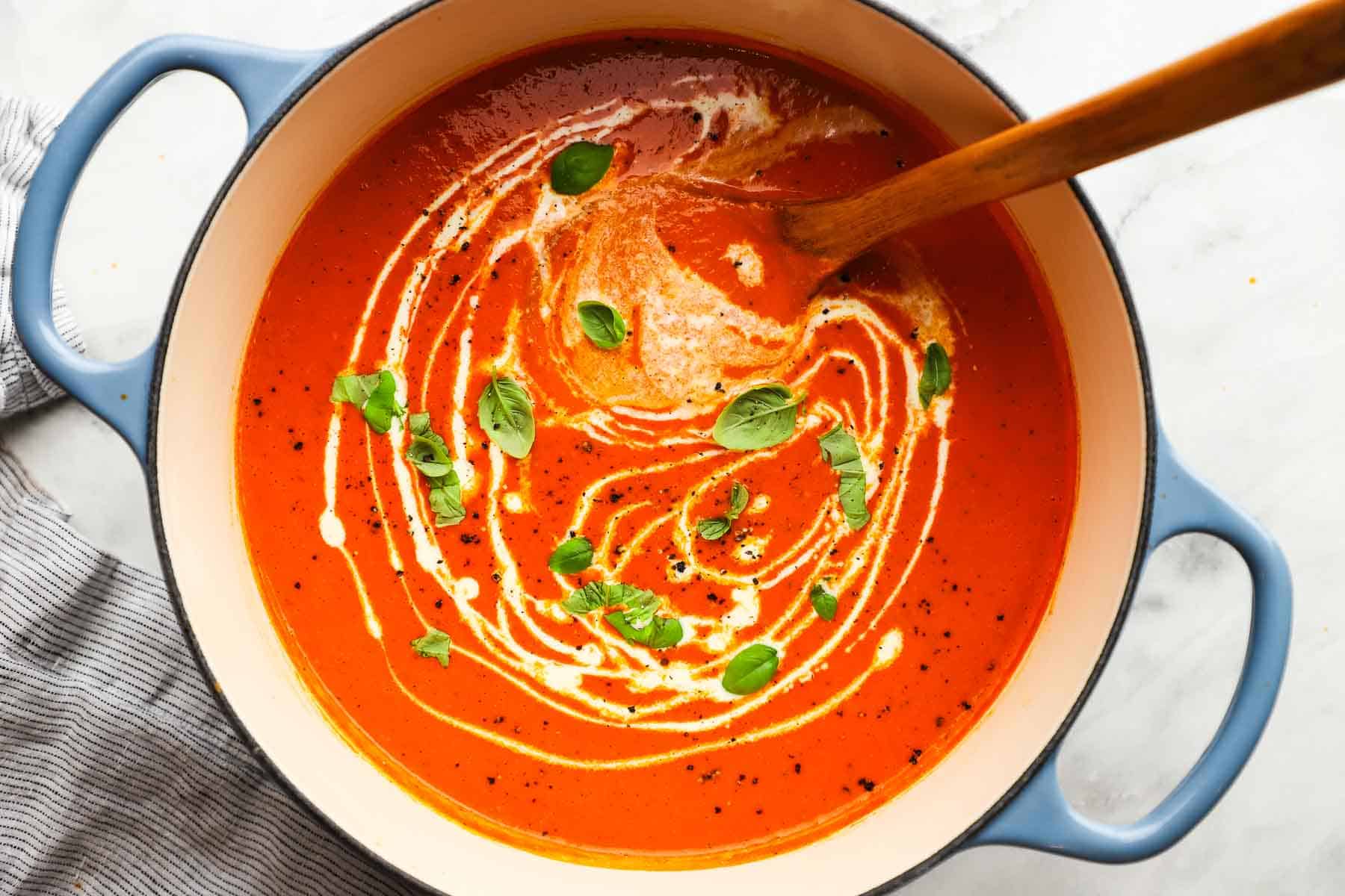 A pot of creamy red pepper soup with a cream swirl and basil garnish.