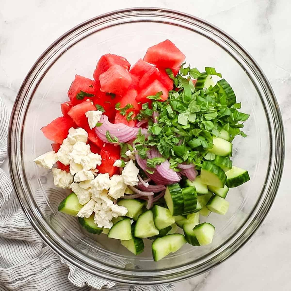 Watermelon, cucumbers, red onion, feta, mint, and basil, all combined in a large serving bowl.