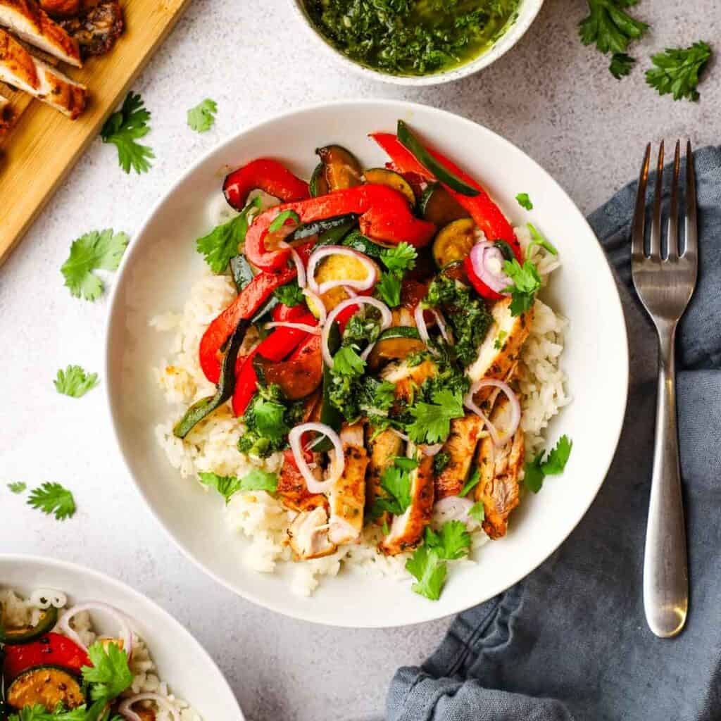 a bowl of rice topped with grilled chicken, veggies, and cimichurri sauce.